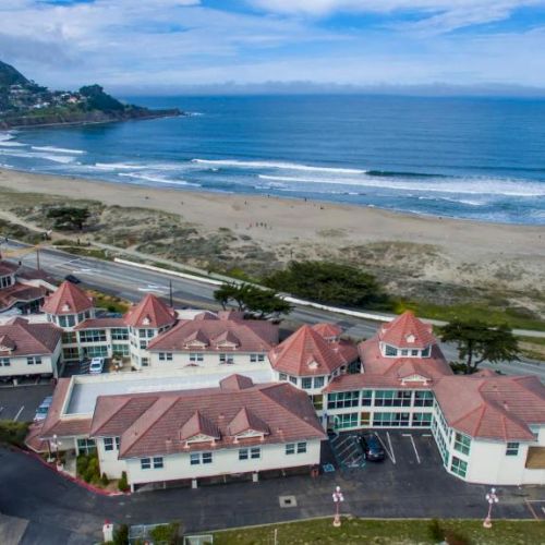 The image shows a coastal hotel with red roofs near a beach and a road, surrounded by greenery and overlooking the ocean under a blue sky.