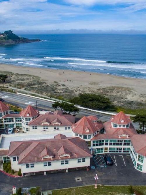 The image shows a coastal hotel with red roofs near a beach and a road, surrounded by greenery and overlooking the ocean under a blue sky.