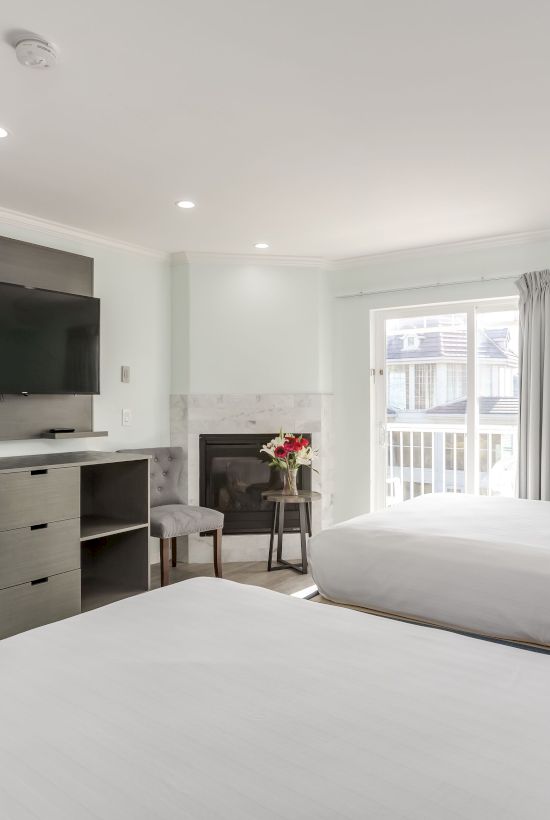 The image shows a modern hotel room with two beds, a desk, a chair, a wall-mounted TV, and a window with curtains letting in natural light.