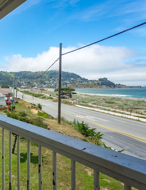 A balcony with white chairs overlooks a coastal road, hillside, and ocean, under a bright blue sky with scattered clouds.