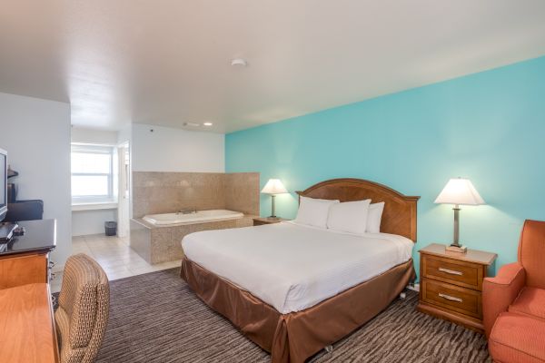 The image shows a hotel room with a large bed, bedside lamps, an armchair, a TV, and a Jacuzzi tub in the corner near the bathroom.