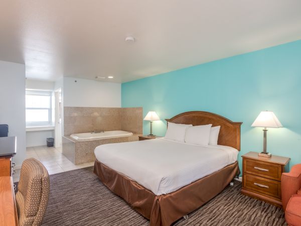 The image shows a hotel room with a large bed, bedside lamps, an armchair, a TV, and a Jacuzzi tub in the corner near the bathroom.