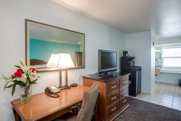 A hotel room with a desk, flowers, lamp, TV, chair, mini fridge, and a mirror on the wall. Bathroom visible in the background.