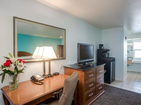 A hotel room with a desk, flowers, lamp, TV, chair, mini fridge, and a mirror on the wall. Bathroom visible in the background.