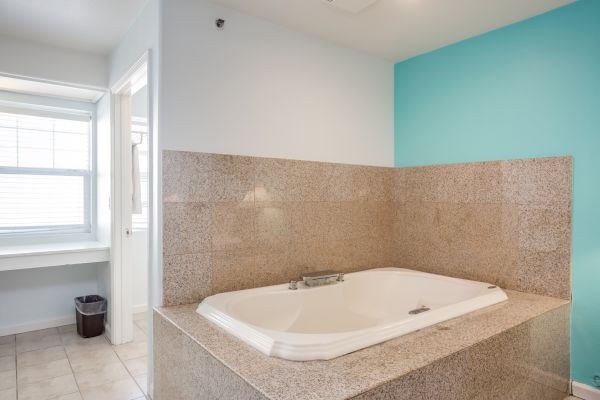 A bathroom with a large, tiled bathtub, turquoise accent wall, window with blinds, tiled floor, and a small trash can in the corner.