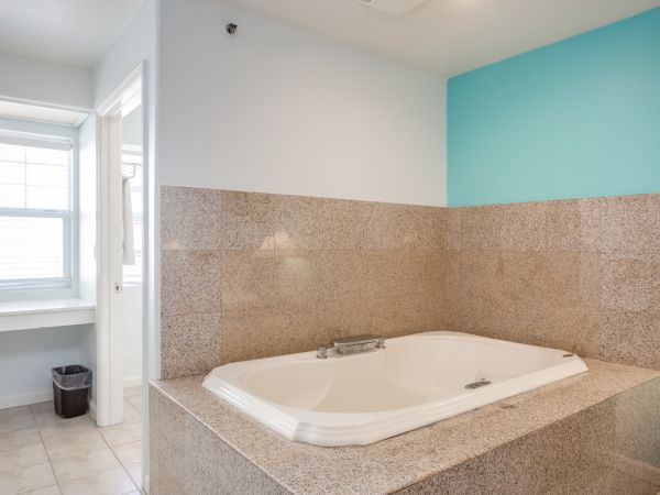 A bathroom with a large, tiled bathtub, turquoise accent wall, window with blinds, tiled floor, and a small trash can in the corner.