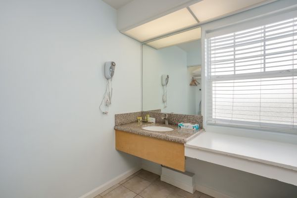 The image shows a bathroom vanity with a mirror, sink, countertop, hairdryer, and a window with blinds. The room has a light, clean design.