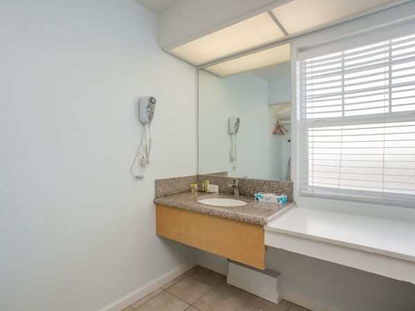 The image shows a bathroom vanity with a mirror, sink, countertop, hairdryer, and a window with blinds. The room has a light, clean design.