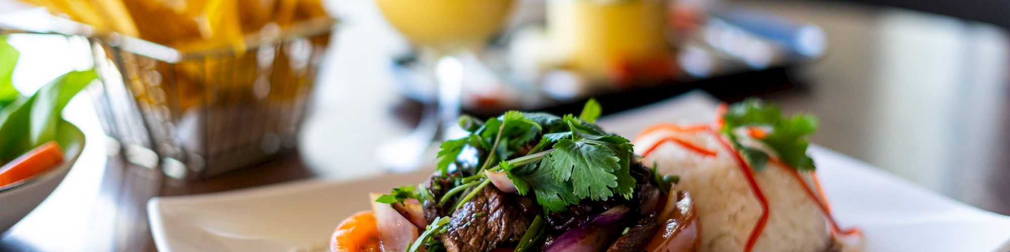 A plate of Lomo Saltado with rice, garnished with cilantro, alongside a basket of fries and two drinks in the background.