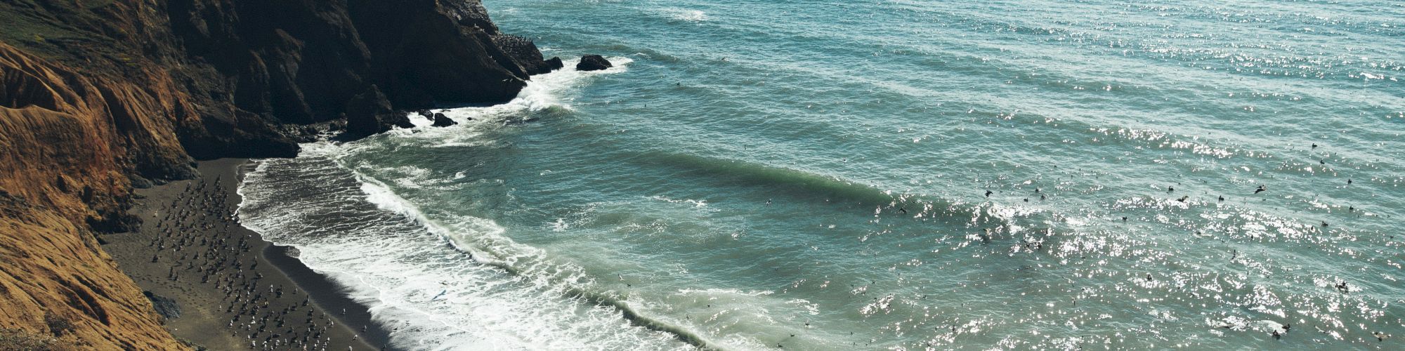 A coastal scene with steep cliffs, a sandy beach, and waves crashing onto the shore under a clear sky, creating a scenic view.