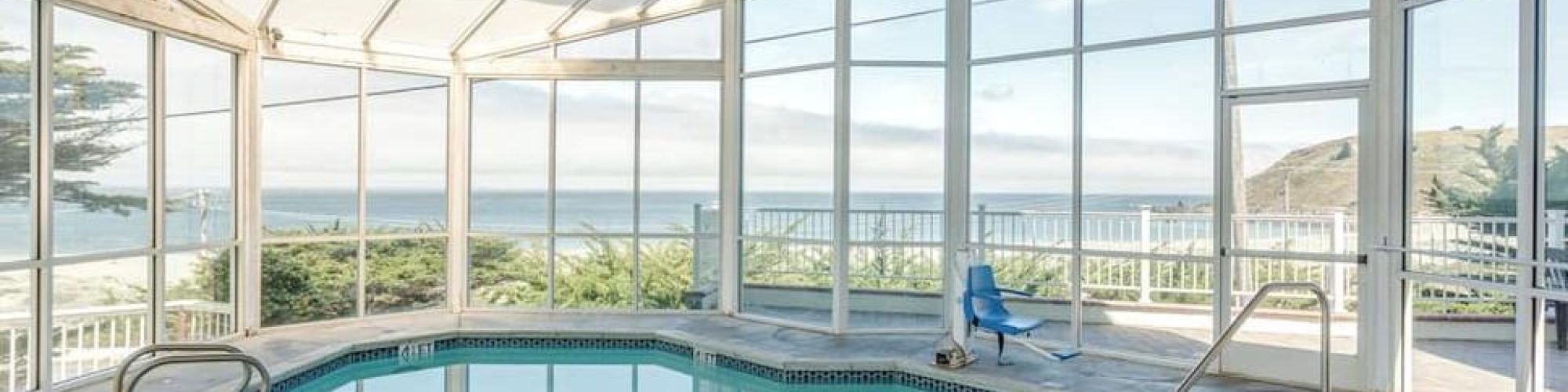 An indoor swimming pool with large windows offers a view of the ocean and surrounding landscape, creating a serene and inviting atmosphere.