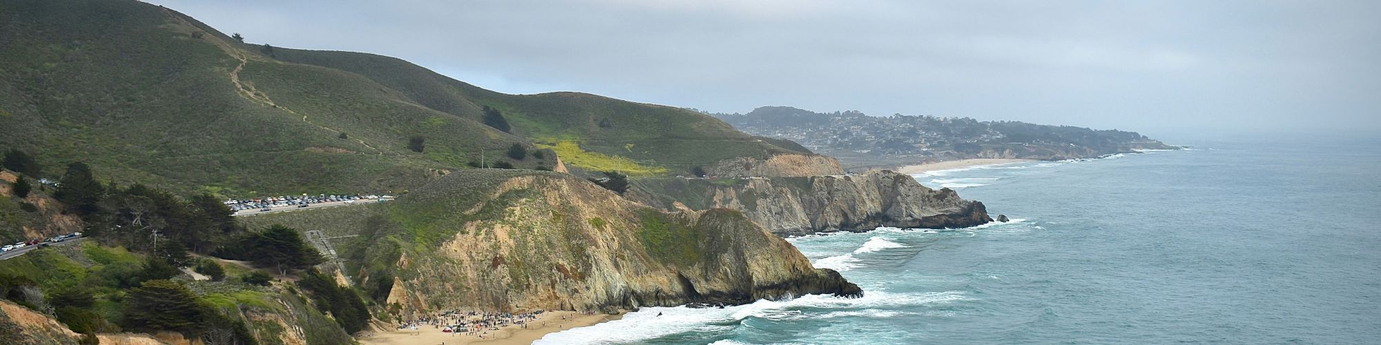 The image shows a coastal scene with waves crashing onto a sandy beach and green hills in the background under a cloudy sky.