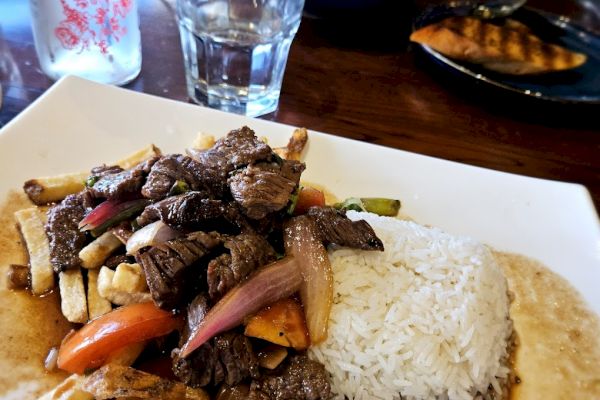 A dish featuring stir-fried beef, onions, and vegetables with rice, likely a Lomo Saltado, alongside a glass of water.