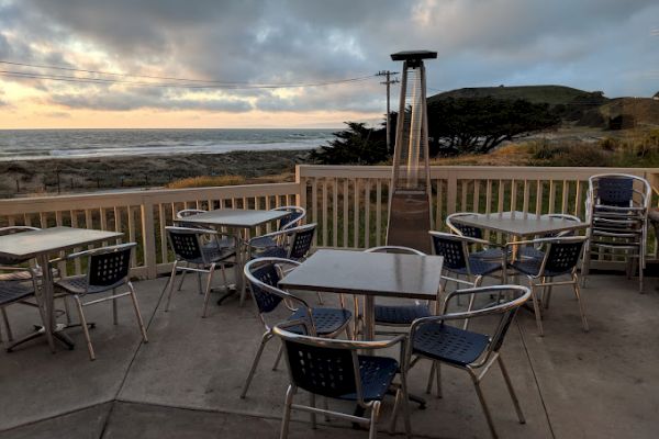 Outdoor patio with metal tables and chairs, overlooking the ocean at sunset. There's a portable heater on the deck.