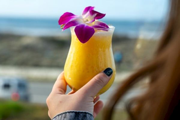 A person holds a yellow drink with a purple flower garnish by the beach, enjoying the ocean view in the background.