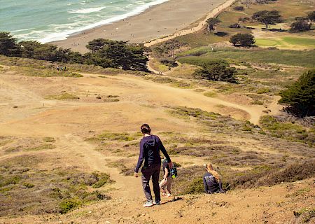 Two people walk down a hill overlooking a scenic coastal landscape with a beach, ocean, and distant town in the background.