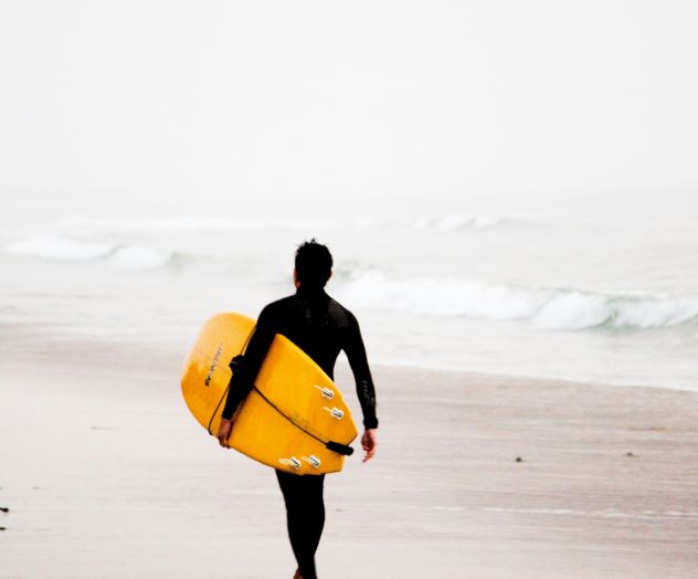 Person walking on a beach towards the ocean, holding a yellow surfboard, with misty weather and scattered rocks on the sand.