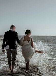 A couple in formal attire walks along the beach, with the woman playfully kicking the water as the waves crash around them.