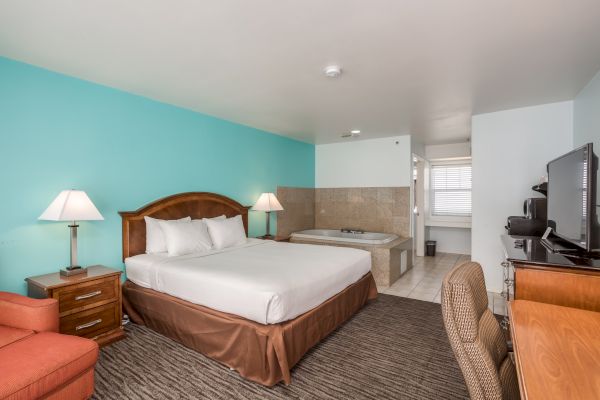 The image shows a hotel room with a bed, two lamps, a TV on a dresser, a chair, and a jacuzzi tub near a tiled bathroom area.