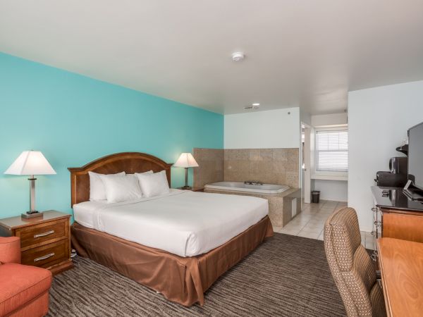 The image shows a hotel room with a bed, two lamps, a TV on a dresser, a chair, and a jacuzzi tub near a tiled bathroom area.