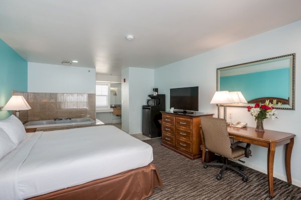 A hotel room with a bed, desk, TV, and mirror. There's a hot tub in the corner, a desk lamp, and a chair. The room has blue and white walls.