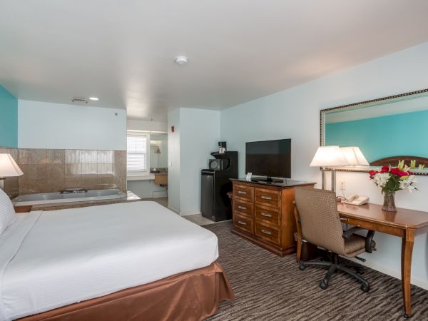 A hotel room with a bed, desk, TV, and mirror. There's a hot tub in the corner, a desk lamp, and a chair. The room has blue and white walls.
