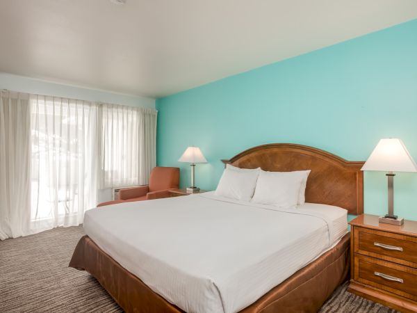 A hotel room features a large bed, two nightstands with lamps, a chair, and a window with white curtains against a turquoise wall.