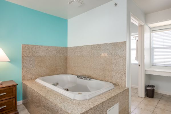 The image shows a bathroom with a jacuzzi tub, beige tile surround, blue accent wall, and a dresser with a lamp, next to a bright window.