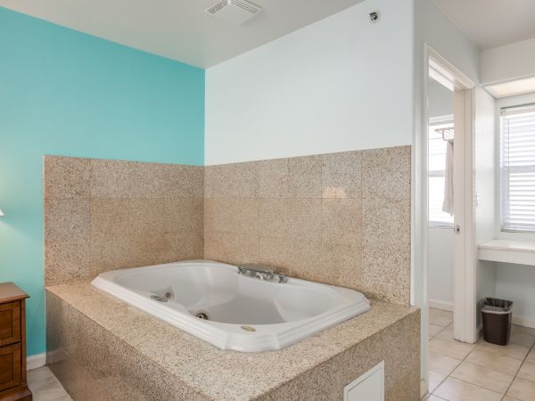 The image shows a bathroom with a jacuzzi tub, beige tile surround, blue accent wall, and a dresser with a lamp, next to a bright window.