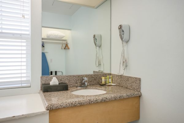 A bathroom with a granite countertop, sink, mirror, wall-mounted hairdryer, toiletries, tissue box, and an ironing board on a shelf.