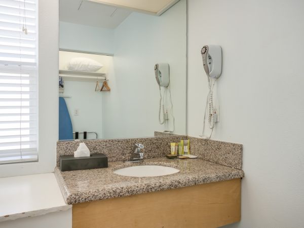 A bathroom with a granite countertop, sink, mirror, wall-mounted hairdryer, toiletries, tissue box, and an ironing board on a shelf.