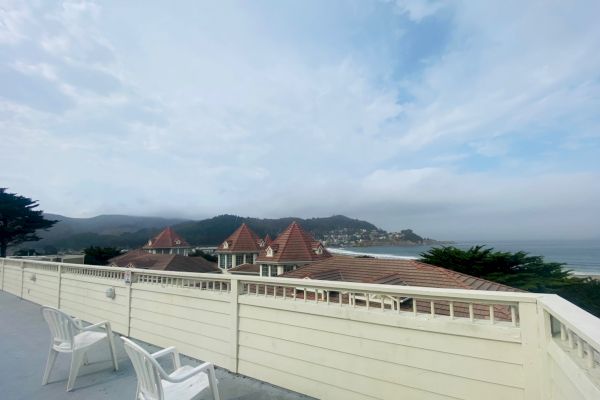 A scenic view of a terrace with white chairs, overlooking rooftops and hills under a cloudy sky, near the coastline.