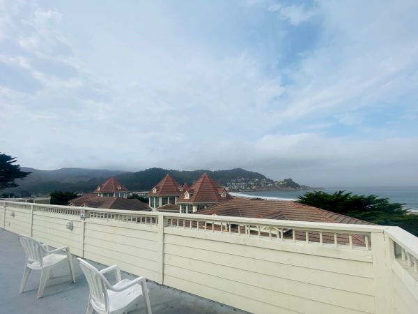 A scenic view of a terrace with white chairs, overlooking rooftops and hills under a cloudy sky, near the coastline.