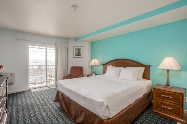 A hotel room with a king-sized bed, teal accent wall, bedside lamps, a chair, and a view through sliding glass doors.