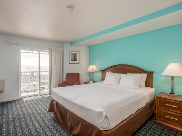 A hotel room with a king-sized bed, teal accent wall, bedside lamps, a chair, and a view through sliding glass doors.