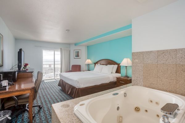 A hotel room with a king-sized bed, a balcony view, a desk with a chair, a flat-screen TV, and a jacuzzi in the foreground.