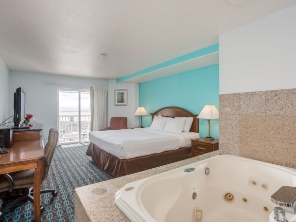A hotel room with a king-sized bed, a balcony view, a desk with a chair, a flat-screen TV, and a jacuzzi in the foreground.