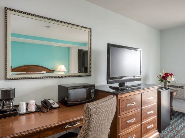 A hotel room desk with a mirror, TV, microwave, coffee maker, chair, lamp, and flowers.