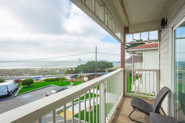 A balcony view with chairs overlooks a road, greenery, and the ocean in the distance under a partly cloudy sky.