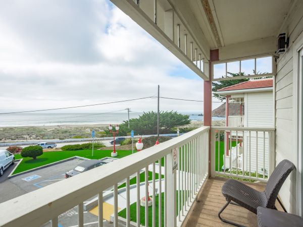 A balcony view with chairs overlooks a road, greenery, and the ocean in the distance under a partly cloudy sky.