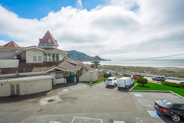 A seaside building with a turret is next to a parking lot with vehicles, overlooking a beach and ocean under a partly cloudy sky.