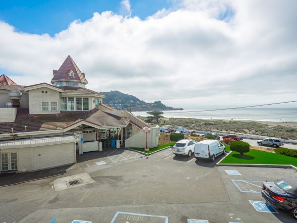 A seaside building with a turret is next to a parking lot with vehicles, overlooking a beach and ocean under a partly cloudy sky.