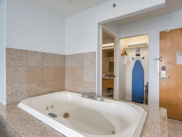 The image shows a bathroom with a whirlpool bathtub, granite tiles, and an ironing board near the entrance of a hotel room.