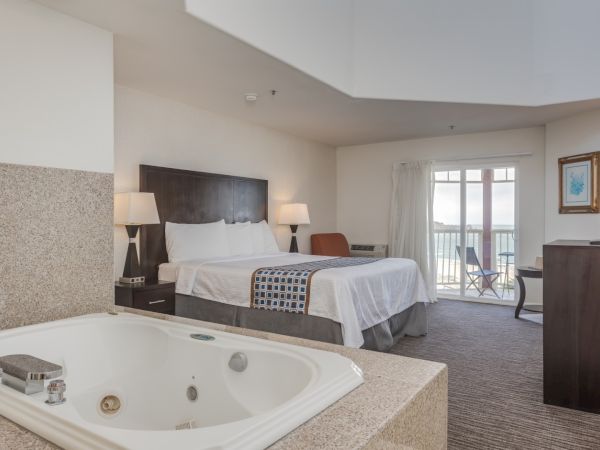 A hotel room with a bed, a jacuzzi tub, a balcony with chairs, TV, and lamps.