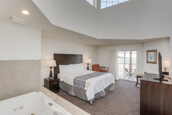 A hotel room with a large bed, a TV, and a balcony. There's a jacuzzi in the corner and a small outdoor seating area is visible.