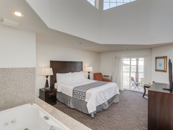 A hotel room with a large bed, a TV, and a balcony. There's a jacuzzi in the corner and a small outdoor seating area is visible.