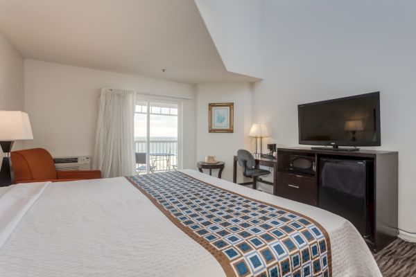 The image shows a hotel room with a bed, TV, desk, chair, and an orange armchair. A balcony with a view is visible through the sliding door.