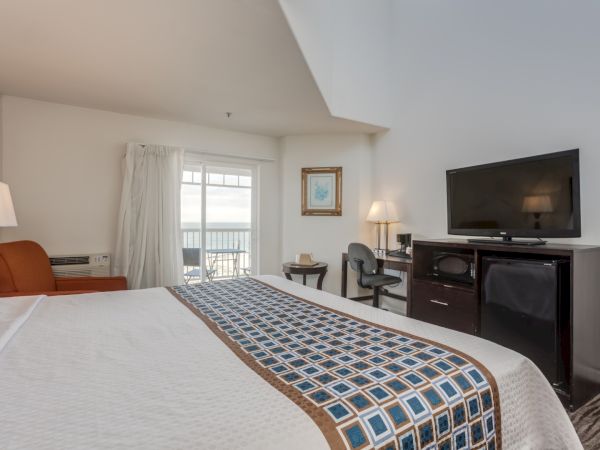 The image shows a hotel room with a bed, TV, desk, chair, and an orange armchair. A balcony with a view is visible through the sliding door.
