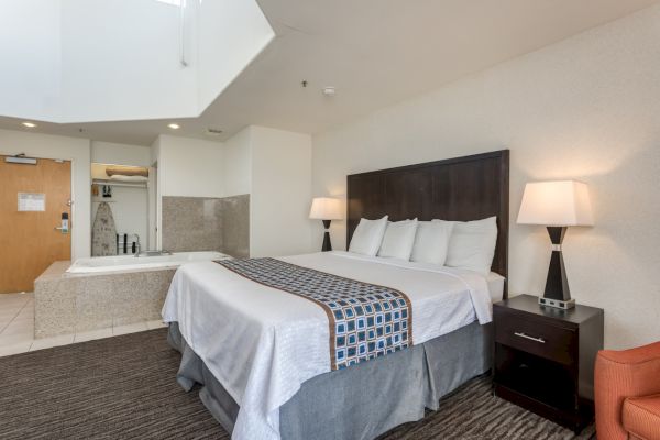 A hotel room with a bed, side table with lamps, carpeted floor, and a corner Jacuzzi tub beside the entrance door.