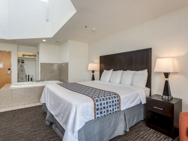 A hotel room with a bed, side table with lamps, carpeted floor, and a corner Jacuzzi tub beside the entrance door.